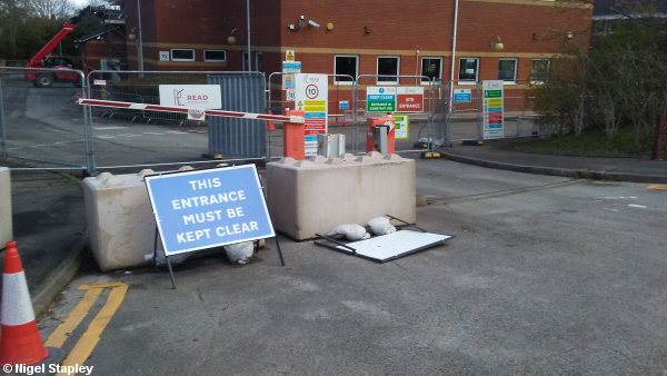 Photo of the entrance to an office car park. Sign says 'This entrance must be kept clear', but entrance is blocked by concrete blocks and wire fences